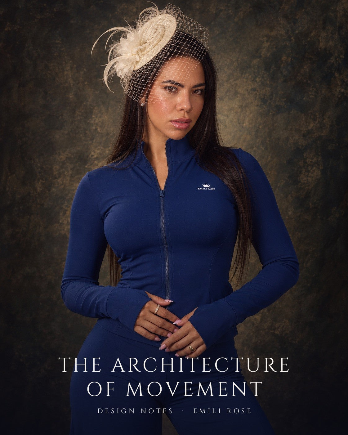 A woman in a navy Emili Rose zip jacket and an ivory feathered fascinator with a birdcage veil, posed against a painted dark backdrop
